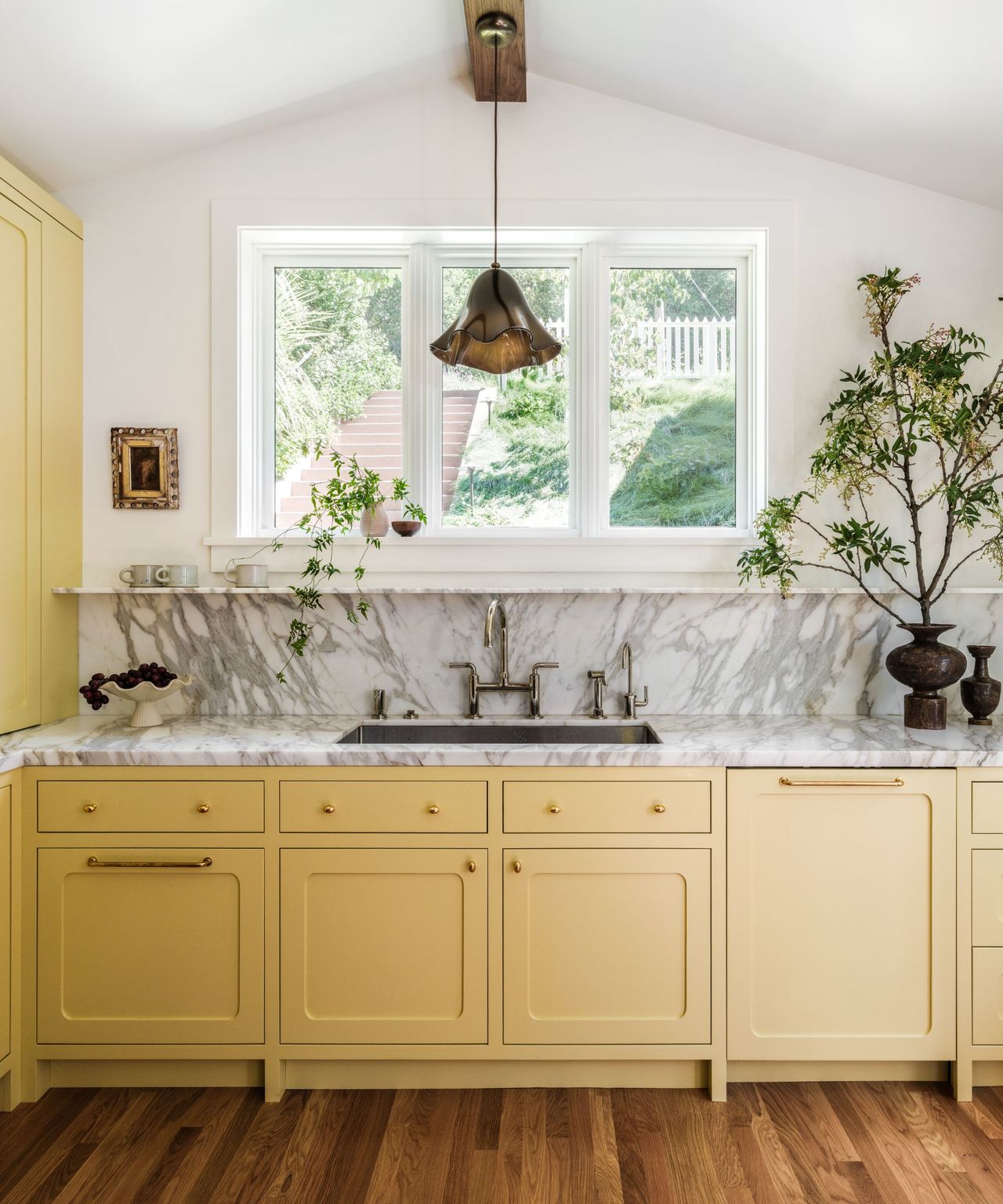 A kitchen with yellow cabinets, a marble countersplash, white walls, brass hardware, and a hand-blown amber glass pendant light