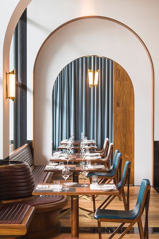 View of a row of tables with tableware and seating at Origin Grill & Bar, Singapore. There are also two gold lights, blue curtains, wood flooring and a window