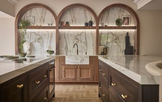 wooden kitchen with arched bespoke shelving, marble worktops and wall panelling and a variety of matching gold hardware on drawers and cabinets