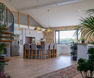 open plan ground floor with white kitchen, large kitchen island with seating, wood and metal spiral staircase and mezzanine level above