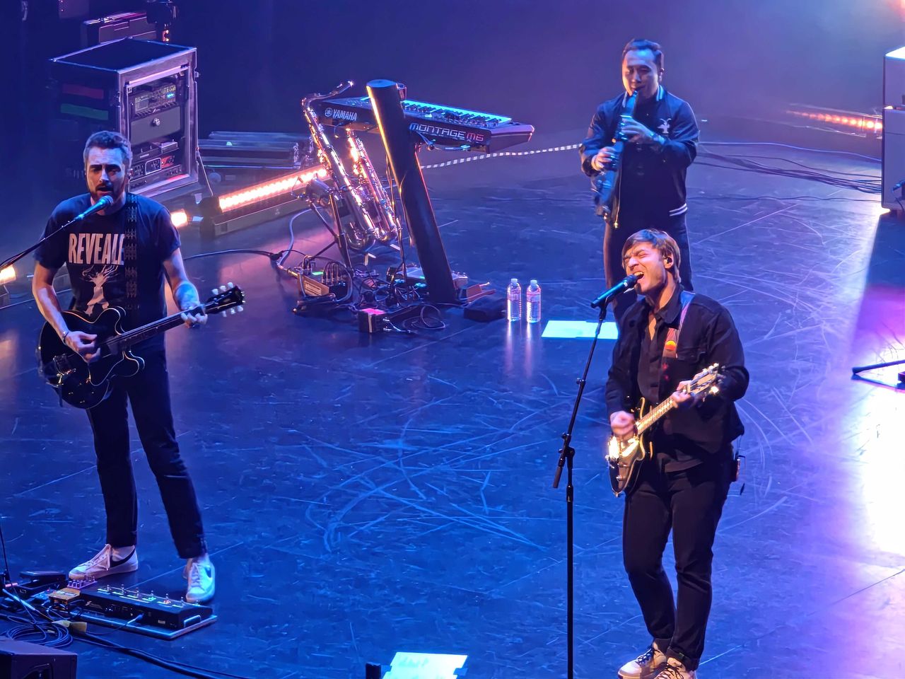 Two male singers with guitars and a third man with a saxophone