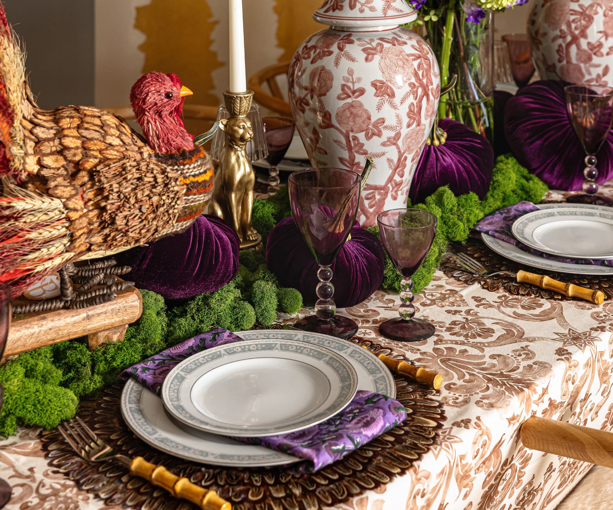 thanksgiving tablescape with a white and brown patterned tablecloth, rattan charger plates, and a large turkey centerpiece with moss and ginger jars