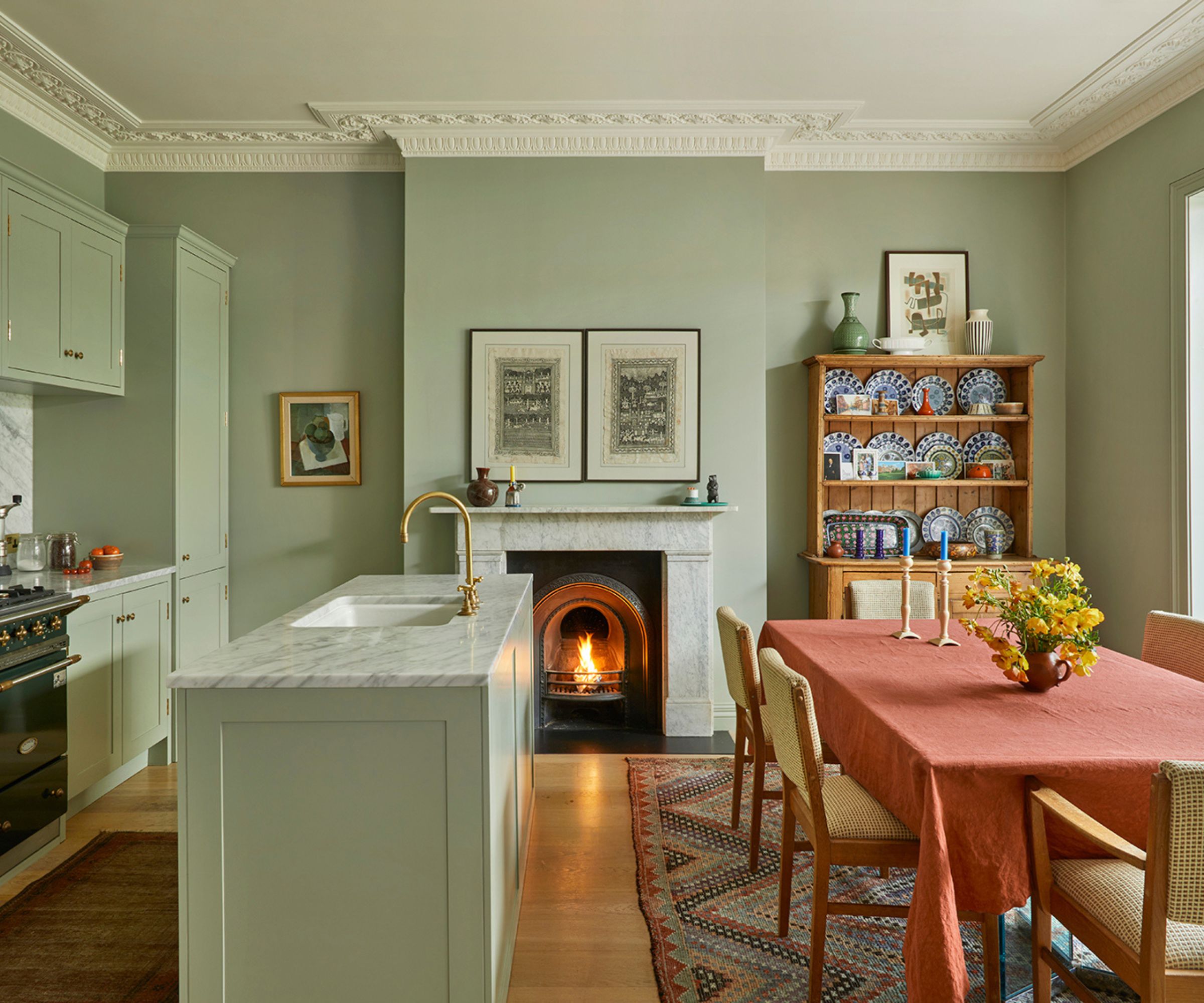 Mint green kitchen with an island, a marble fireplace at the centre with a pair of vintage prints above, a dresser filled with china, and a dining table with an orange linen tablecloth draped on top.