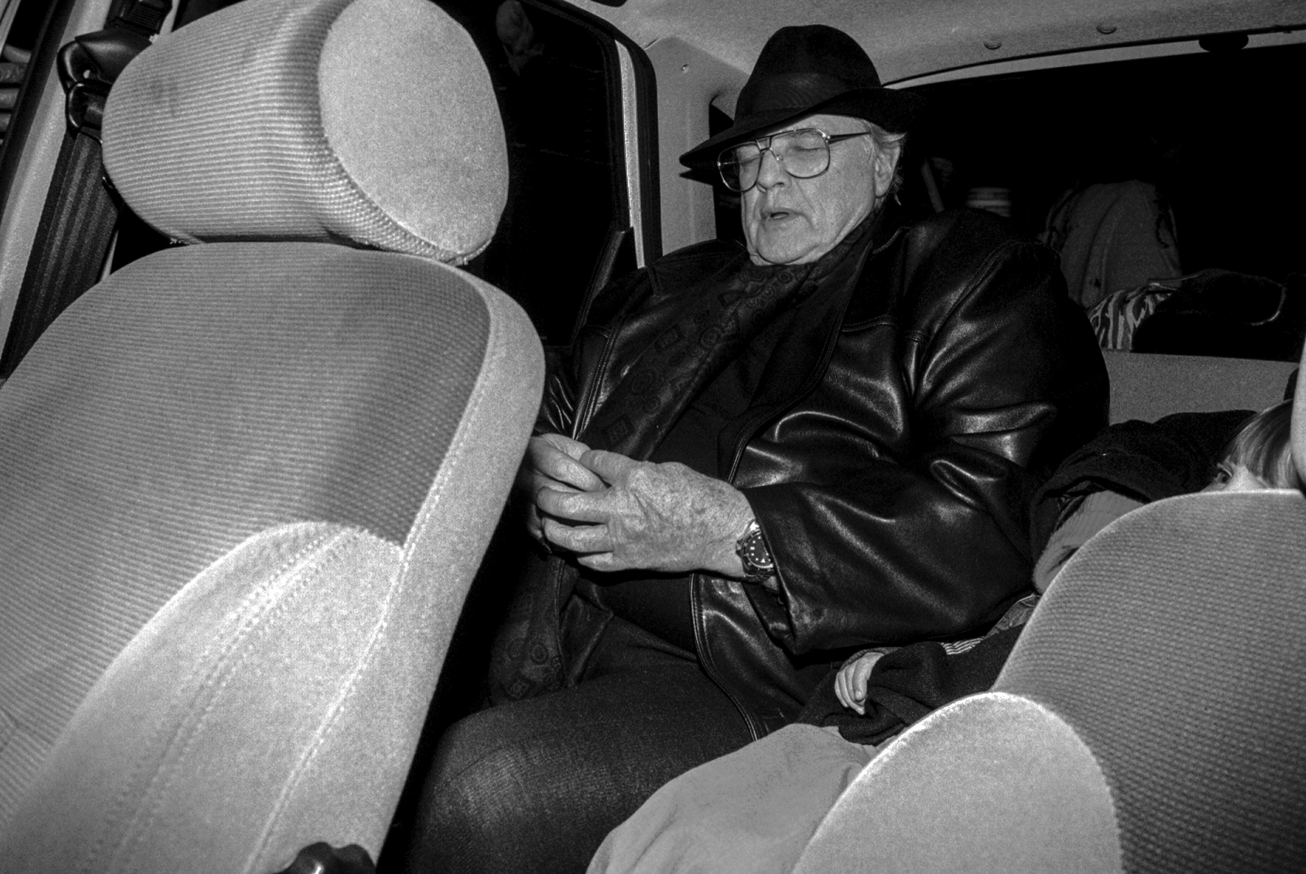 Black and white photo of an older Marlon Brando in a fedora, glasses and leather jacket, looking down as he sits in the back of a taxi at night.