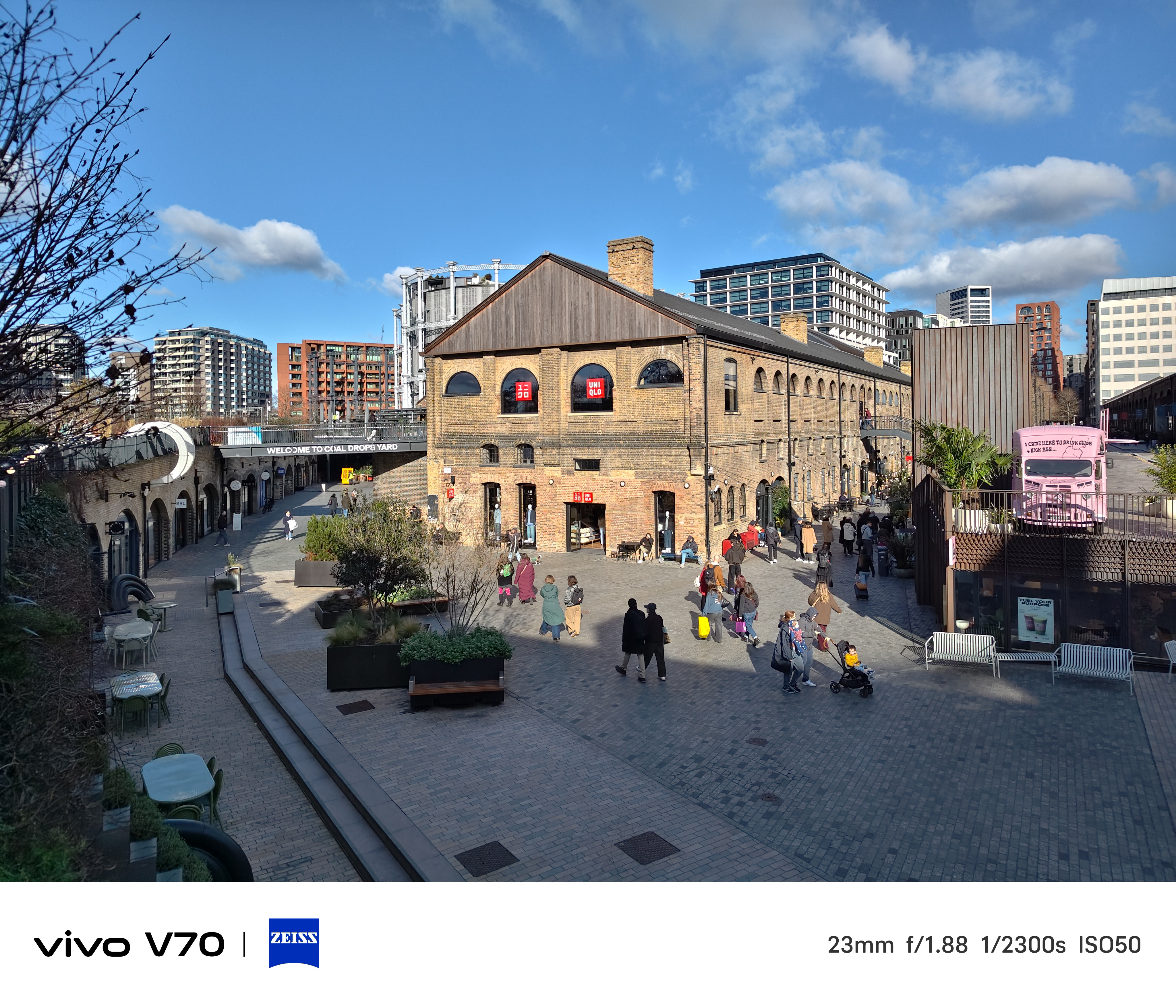 Sunny wide shot of Coal Drops Yard courtyard with shoppers walking between historic brick buildings.