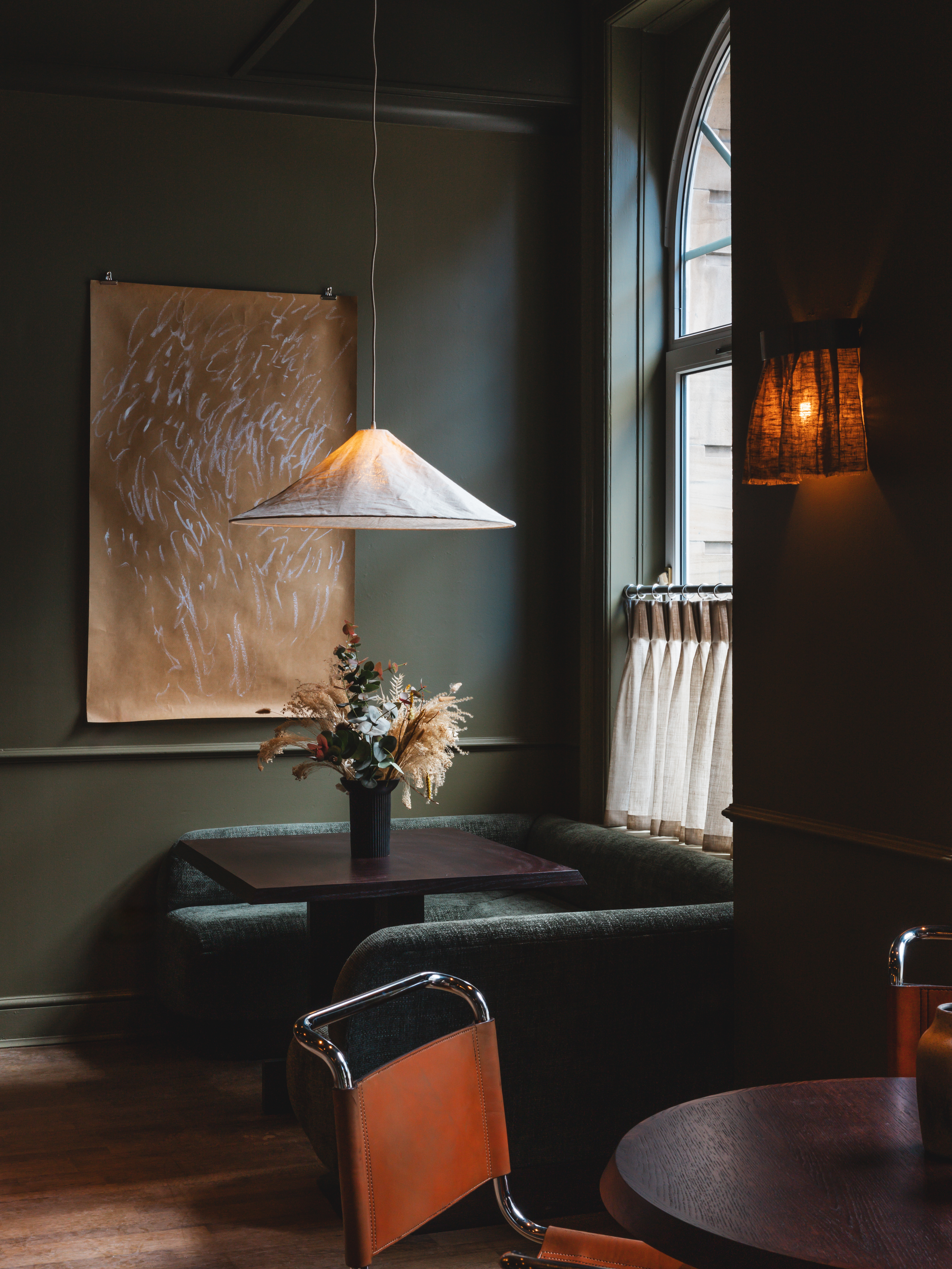 A dark, muted green-tinted hotel lounge with velvety chubby seating in similarly toned nuances, wall art, and wooden furniture.