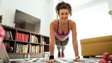 A woman performs a push-up at home in a living room. She is on her hands and toes, her body held in a straight line. Behind her we see a television standing on a low bookshelf.