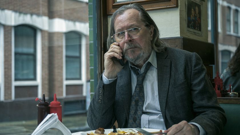 A man (Gary Oldman playing the character of Jackson Lamb) sitting at a table by the window in a cafe. He has a full English breakfast on a plate in front of him and is holding a mobile phone to his right ear.