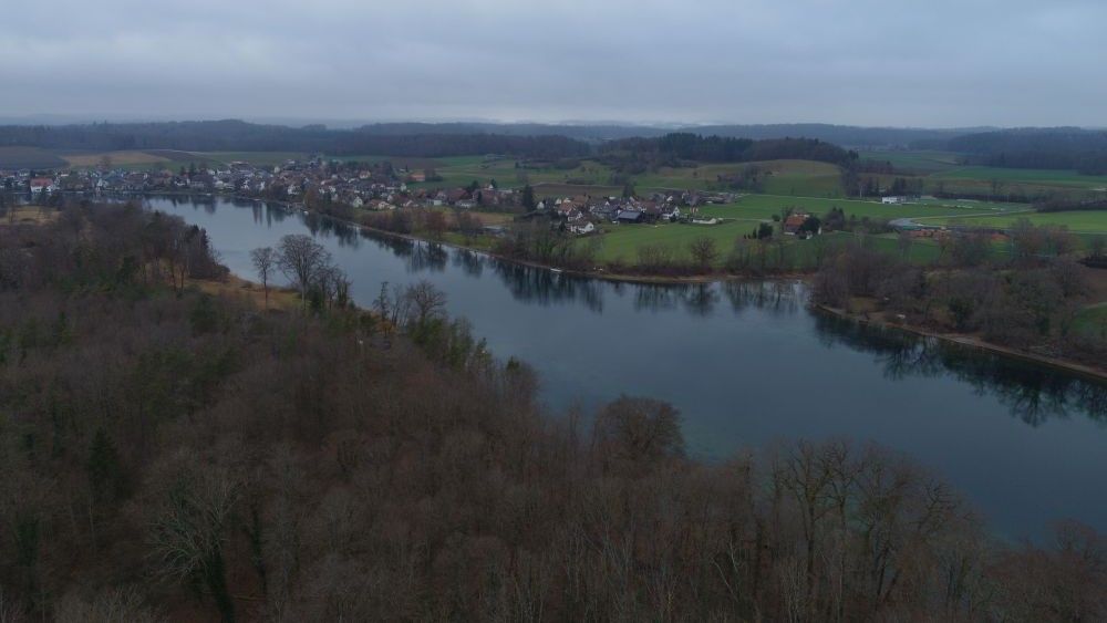 A drone shot of the area to the north of the watchtower. There is a wide river running from the top left to the bottom right of the image. On the bank closest to us are lots of trees (without their leaves). On the far side of the bank there is a small town.