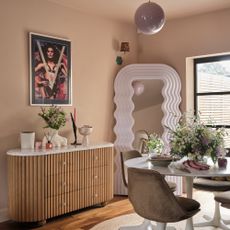 A pink-painted dining area with a white round dining table, a reede console table and a wavy full-length mirror in the corner