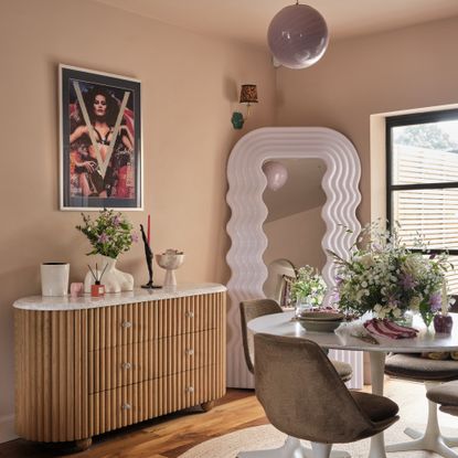 A pink-painted dining area with a white round dining table, a reede console table and a wavy full-length mirror in the corner