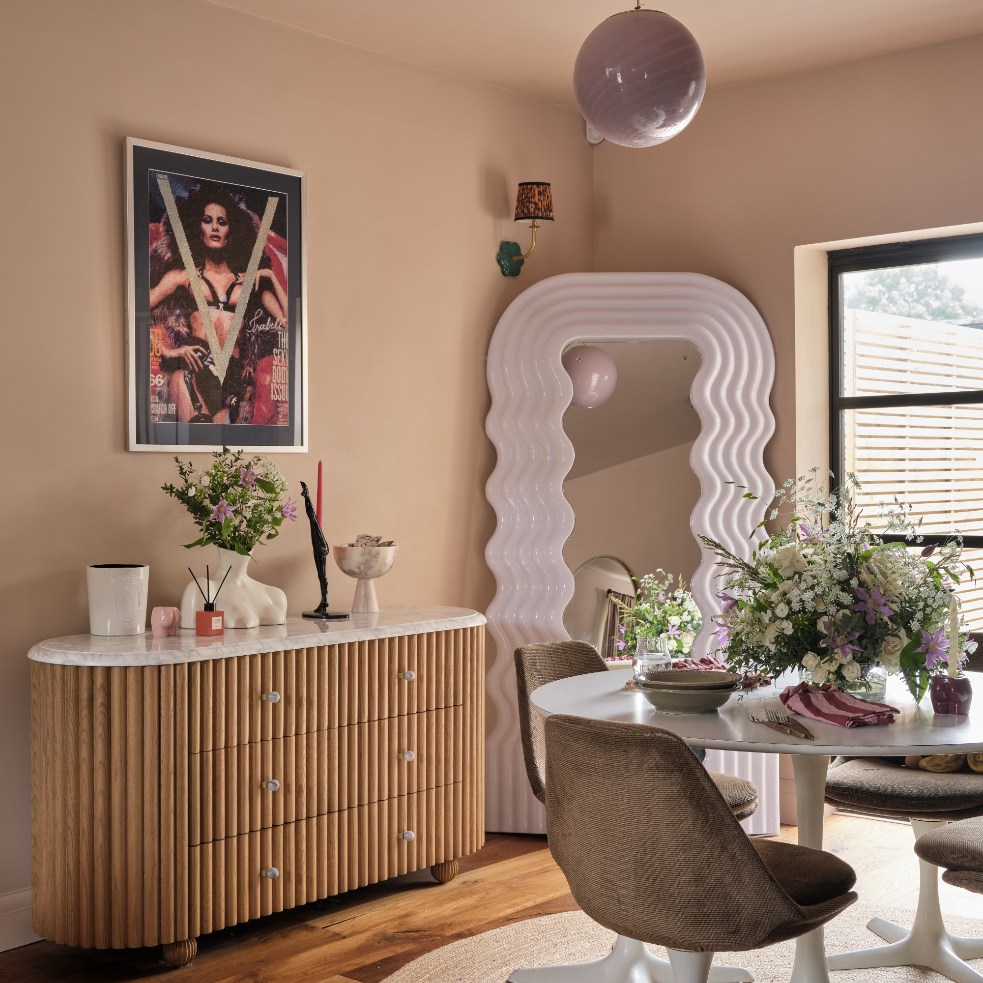 A pink-painted dining area with a white round dining table, a reede console table and a wavy full-length mirror in the corner
