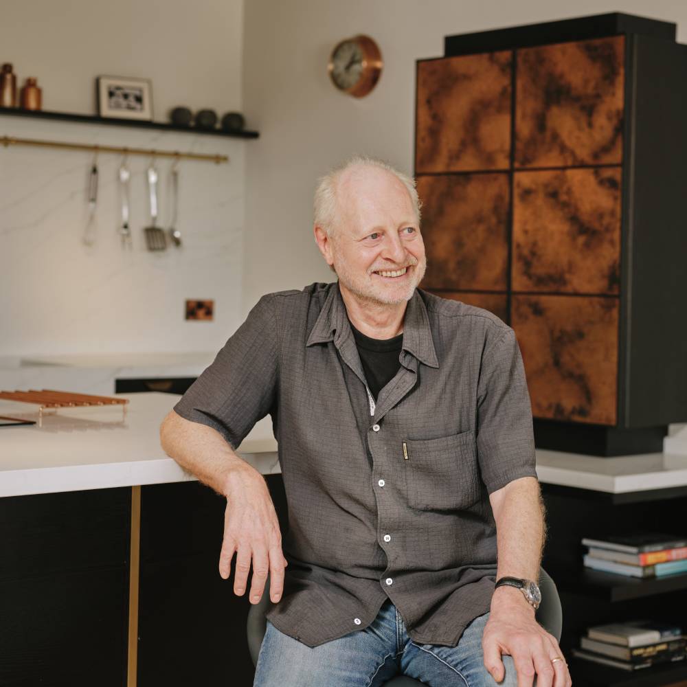 male with short white hair wearing grey shirt and jeans sat at kitchen island