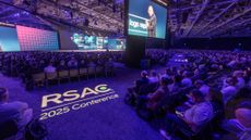 A wide-angle shot of the keynote hall at RSAC Conference 2025, held at the Moscone Center in San Francisco. The RSAC Conference 2025 logo is projected on the floor and the whole room, filled with attendees, is bathed in purple light.