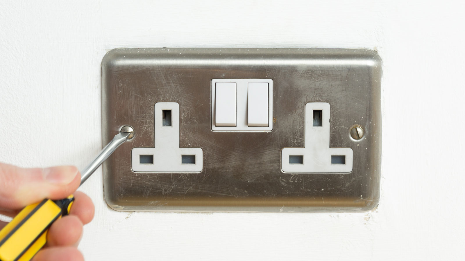 Aluminium double electric socket on white wall with yellow handled screwdriver undoing screw.