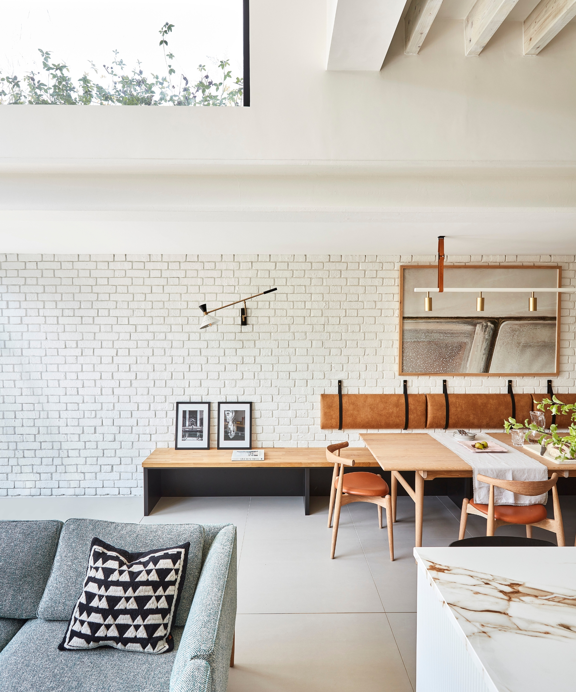 Open-plan living space with painted brick wall, wooden dining table, tan leather bench, high beamed ceiling.