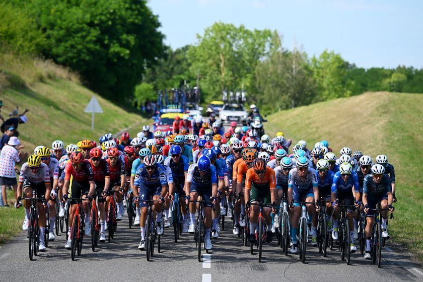 The Tour de France peloton in 2024 riding on a narrow road between two grassy berms with a blue skie overhead