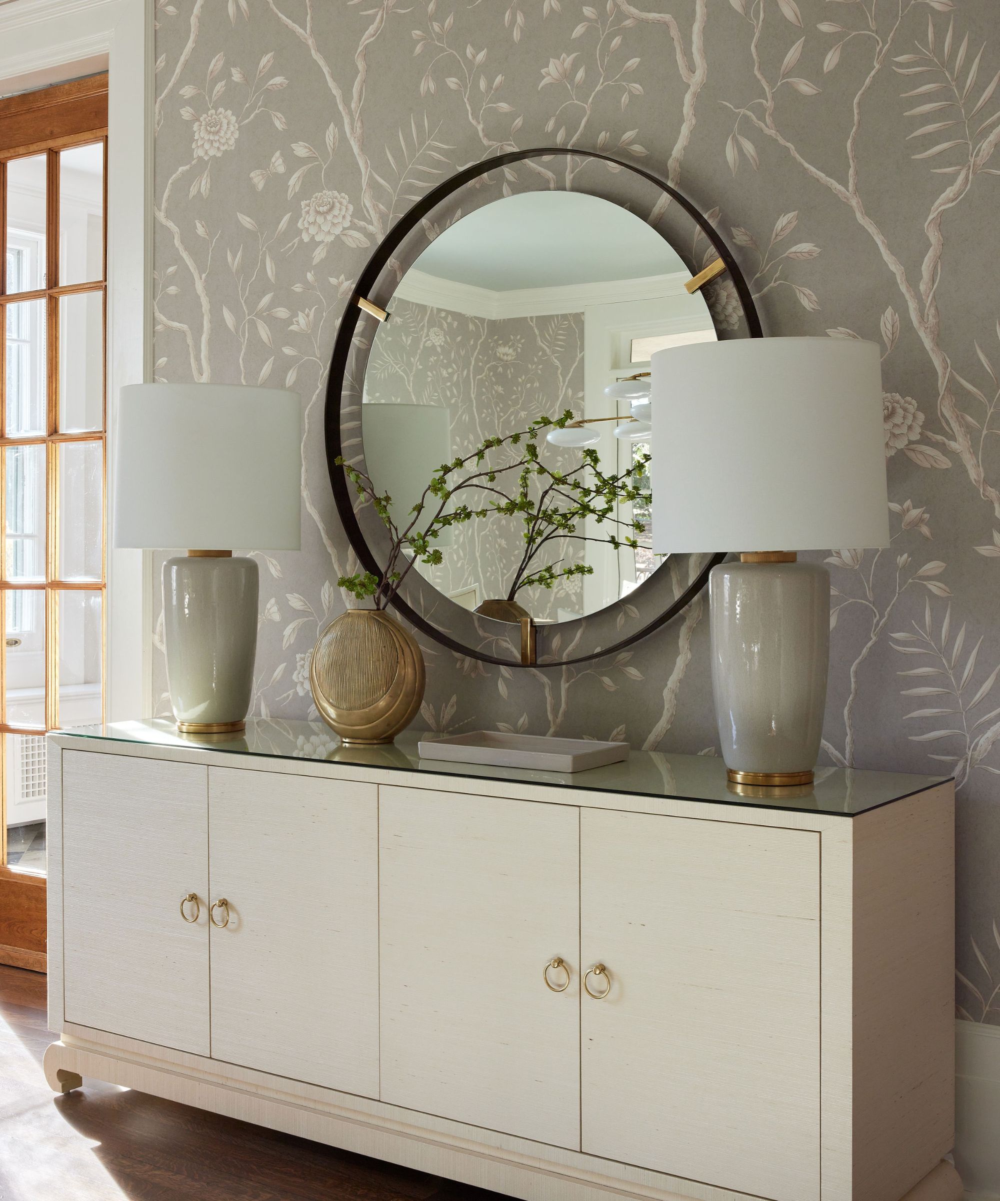A corner of a transitional living room with gray and white forest-motif wallpaper, a creamy white side cabinet with two gray table lamps, a vase of foliage, and a modern round mirror on the wall.