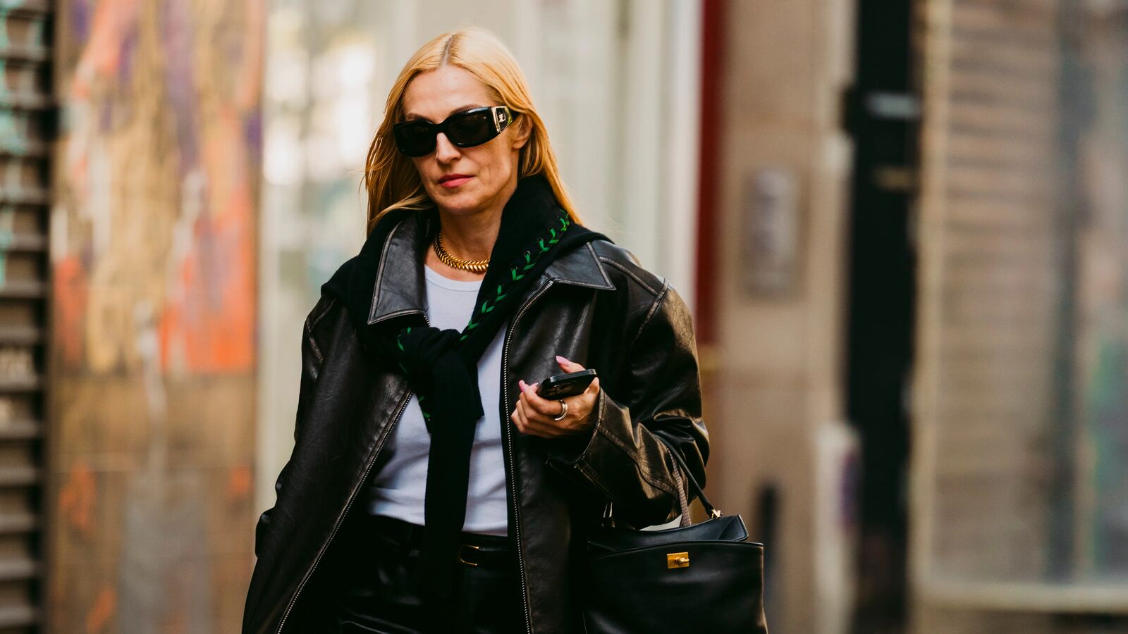 Woman wears black leather jacket, skirt, and handbag. She wears a lighter blue or white top with a black sweater tied around her neck. She has pointed toe heels with a lot of metal hardware on them. 