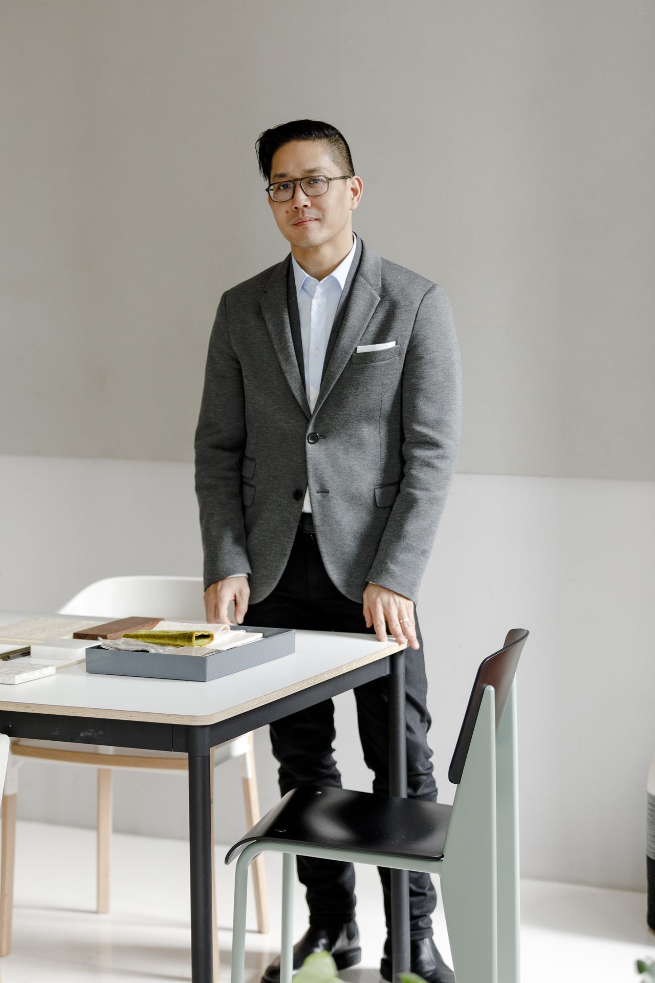A young man dressed in a grey tailored jacket, light blue shirt, and dark trousers stands behind a white desk featuring the prototype of an architectural model.