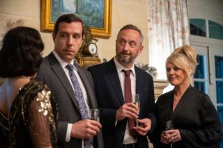 Caron (Alex Gaumond), Dom (Steve Edge) and Jean (Sally Lindsay) attend a murder mystery evening. Caron and Dom are wearing suits, Jean is wearing a black evening dress. They are talking to one of the actors from the murder mystery cast, who has her back to the camera and is dressed in a 1920s-style outfit.