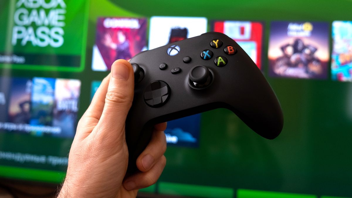 An Xbox Series X controller held in front of a TV showing the console&#039;s dashboard