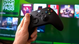 An Xbox Series X controller held in front of a TV showing the console's dashboard