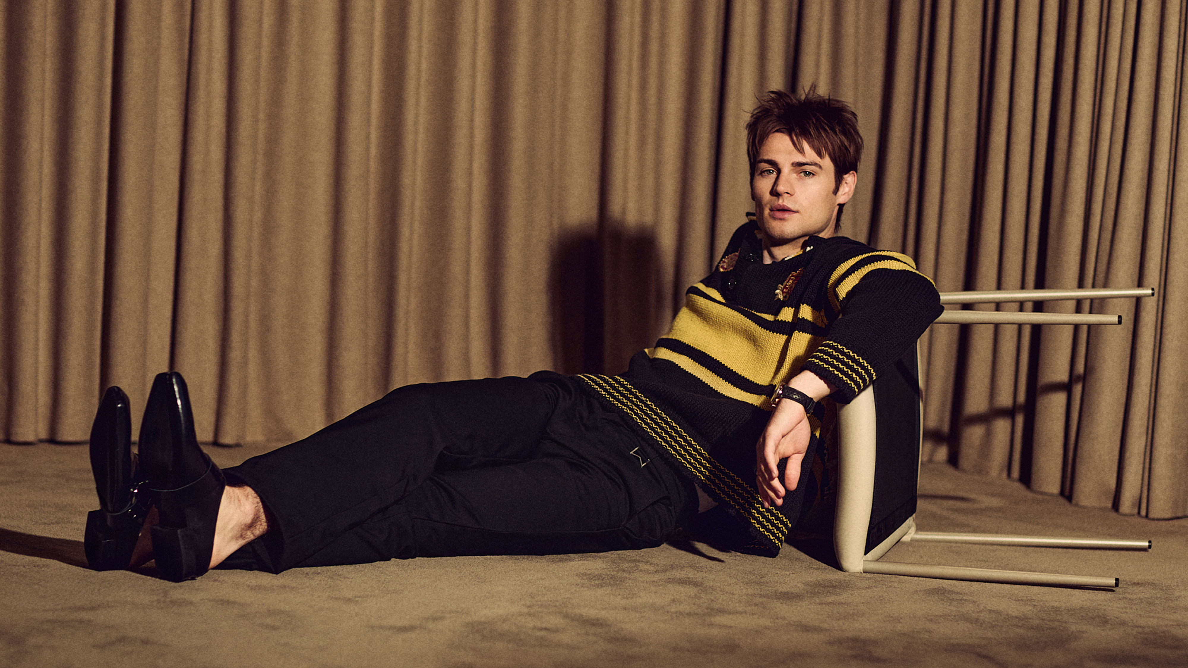 My Life With the Walter Boys actor Noah LaLonde for Who What Wear. He is sitting on the ground in front of a beige curtain leaning against a chair that is tipped on its side. He is wearing a yellow and black sweater with black pants and shoes.
