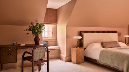A bedroom with sloped ceilings, color-drenched in a soft shade of pink. A neutral carpet, a low-profile bed with neutral bedding, a wooden desk and chair, and a burl wood nightstand.