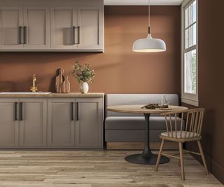 Warm, modern kitchen nook featuring a sleek grey banquette, round wooden dining table with black pedestal base, wooden chair, and soft pendant lighting against terracotta walls.