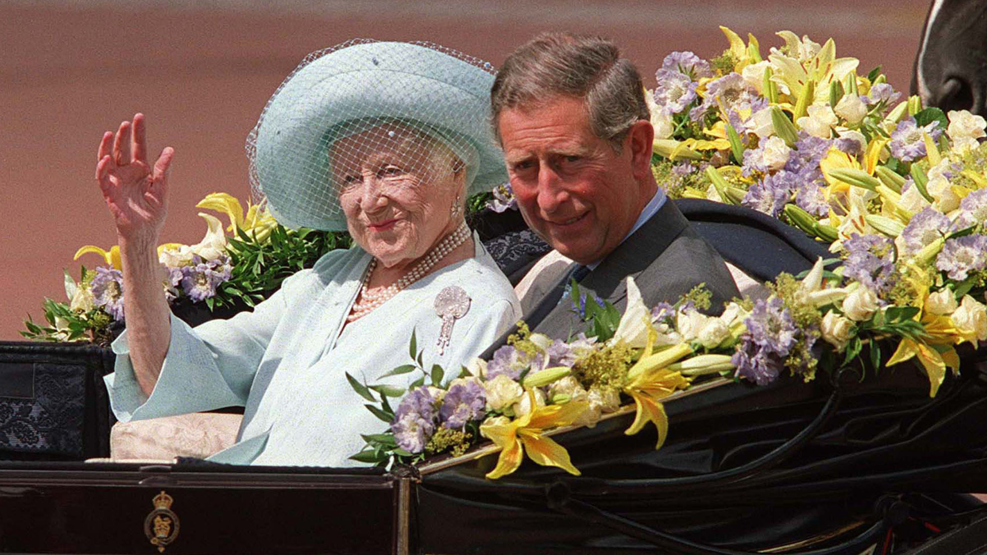 The Queen Mother wearing a blue hat and coat waving from a carriage with King Charles