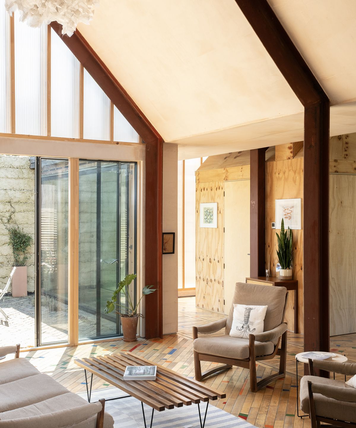 A modern living area in a slef build with pitched ceilings and glazed gable with glass sliding door letting in lots of natural light. The furniture has a midcentury modern aesthetic and the materials on display are natural