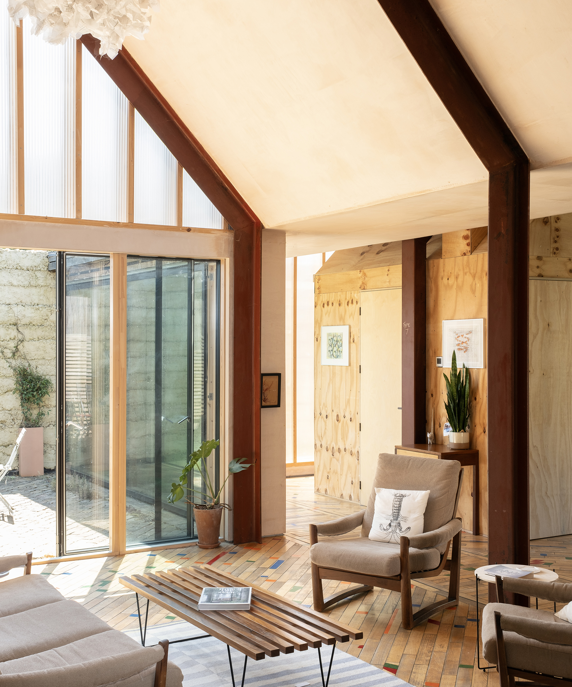A modern living area in a slef build with pitched ceilings and glazed gable with glass sliding door letting in lots of natural light. The furniture has a midcentury modern aesthetic and the materials on display are natural