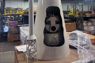 A large-scale cutaway model of an Apollo spacecraft as it would be mounted atop the Saturn V rocket sits atop a table in a warehouse