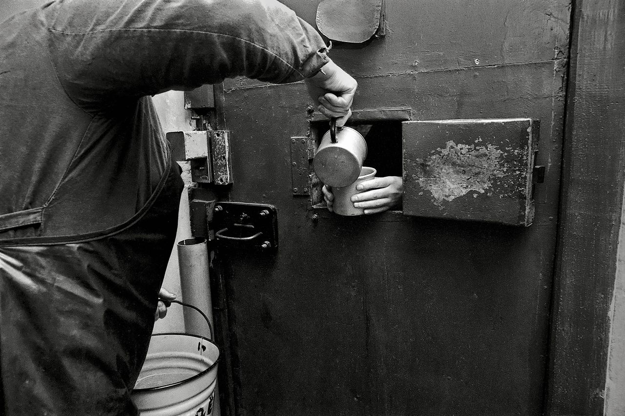 Black and white photograph of someone pouring liquid into a cup held by a prisoner through a small opening in a door