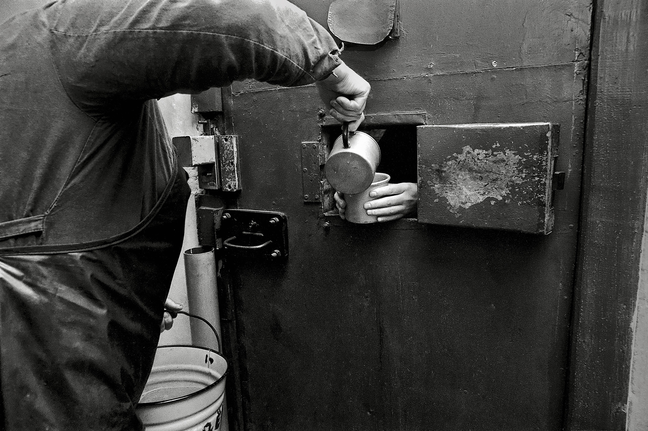 Black and white photograph of someone pouring liquid into a cup held by a prisoner through a small opening in a door