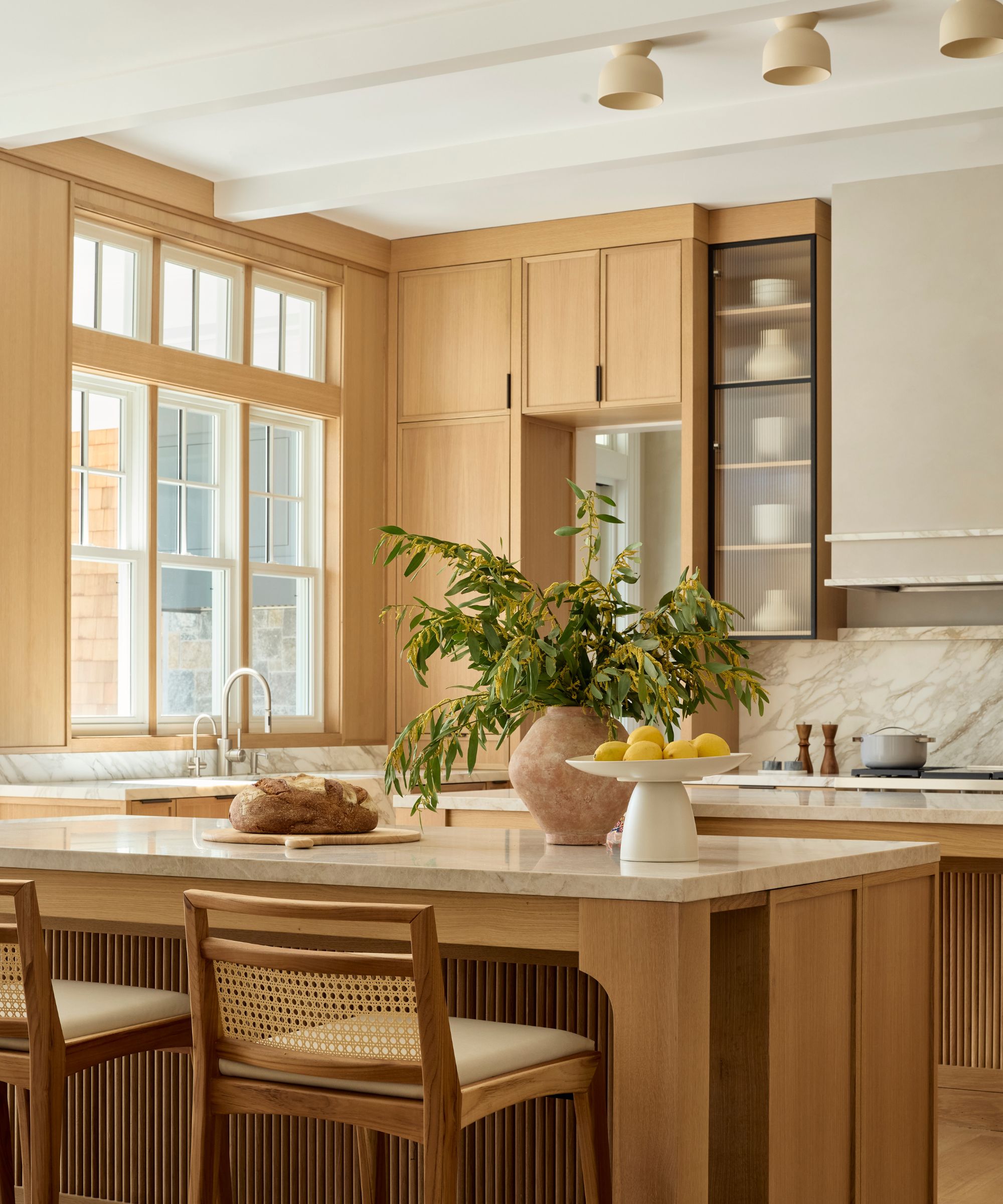 Warm, light wood kitchen island with marble countertop and cane barstools, featuring a large clay vase filled with leafy green branches in the background.