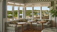 A bright, corner wrap-around booth with pale green cushions and patterned throw pillows, surrounded by large windows looking out onto an outdoor patio with umbrellas and greenery.
