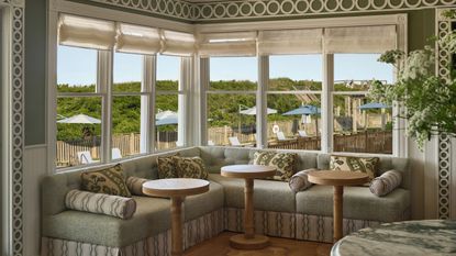 A bright, corner wrap-around booth with pale green cushions and patterned throw pillows, surrounded by large windows looking out onto an outdoor patio with umbrellas and greenery.