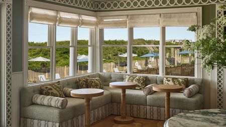 A bright, corner wrap-around booth with pale green cushions and patterned throw pillows, surrounded by large windows looking out onto an outdoor patio with umbrellas and greenery.