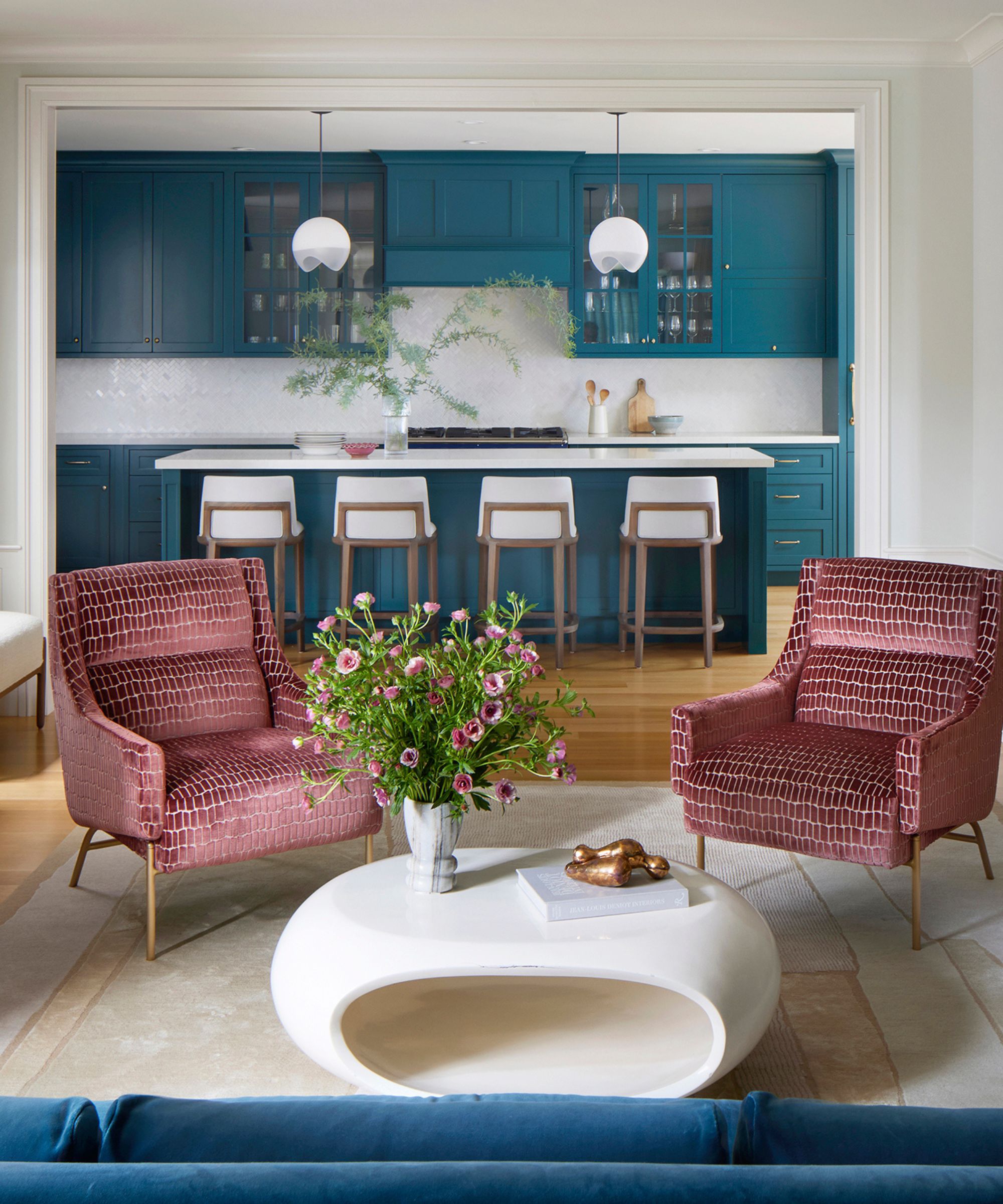 Photograph of sitting area with two red armchairs, curved white coffee table with flowers and paperweight on marble floor. Pictured in background is a blue kitchen with four white stools, two hanging lights and blue cabinets.