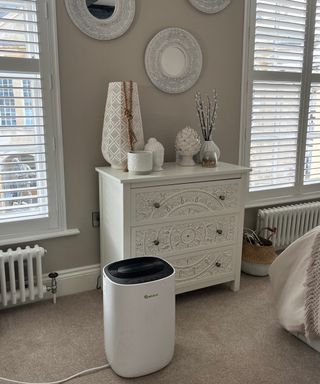 dehumidifier in bedroom with cream carpet, cream chest of drawers in between windows with white shutters