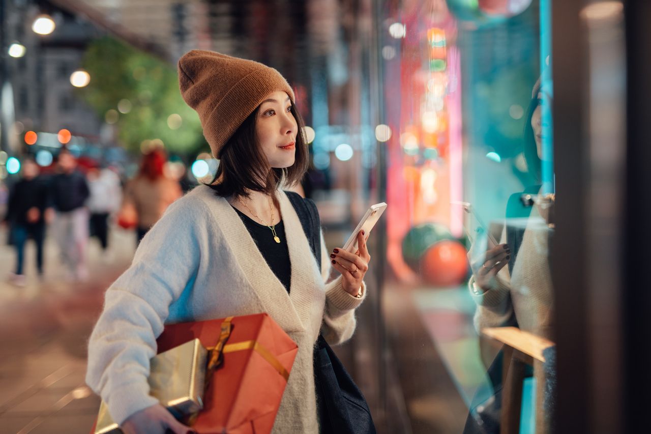 young Asian woman holding smart phone and Christmas gifts, looking at shop window on shopping street