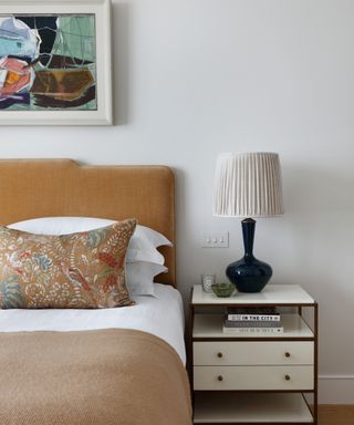 neutral bedroom with gold velvet headboard, patterned cushion, beige throw, artwork and white bedside chest