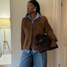 Fashion influencer Nnenna wearing wide-leg jeans, silver statement earrings, a striped button-down shirt, a brown suede jacket, and carrying a suede bag.