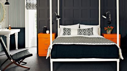 An example of dark bedrooms showing a bedroom with black panelled walls, white four poster bed and orange side tables