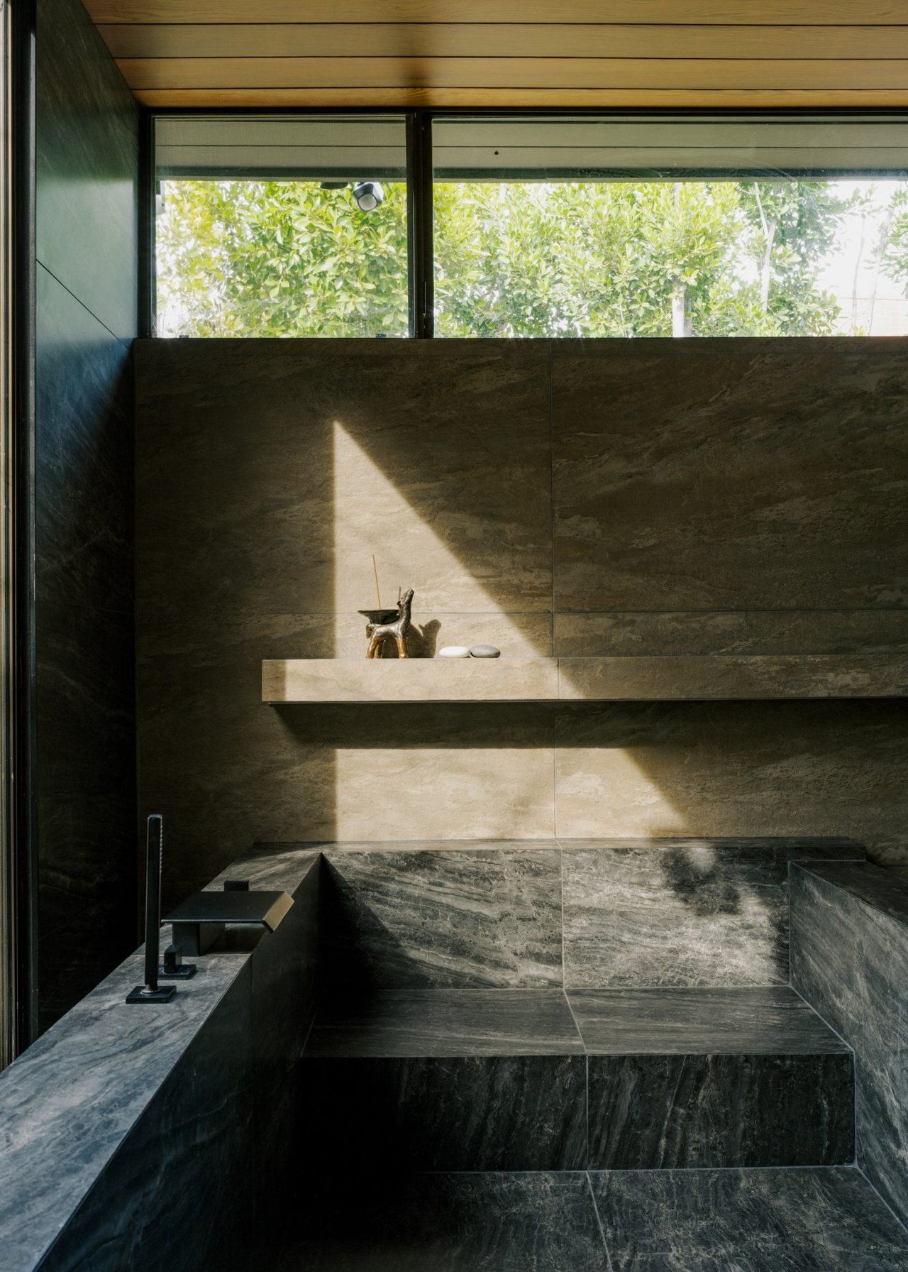 A bathroom with a dark stone tub and a beige stone ledge with light peeking in