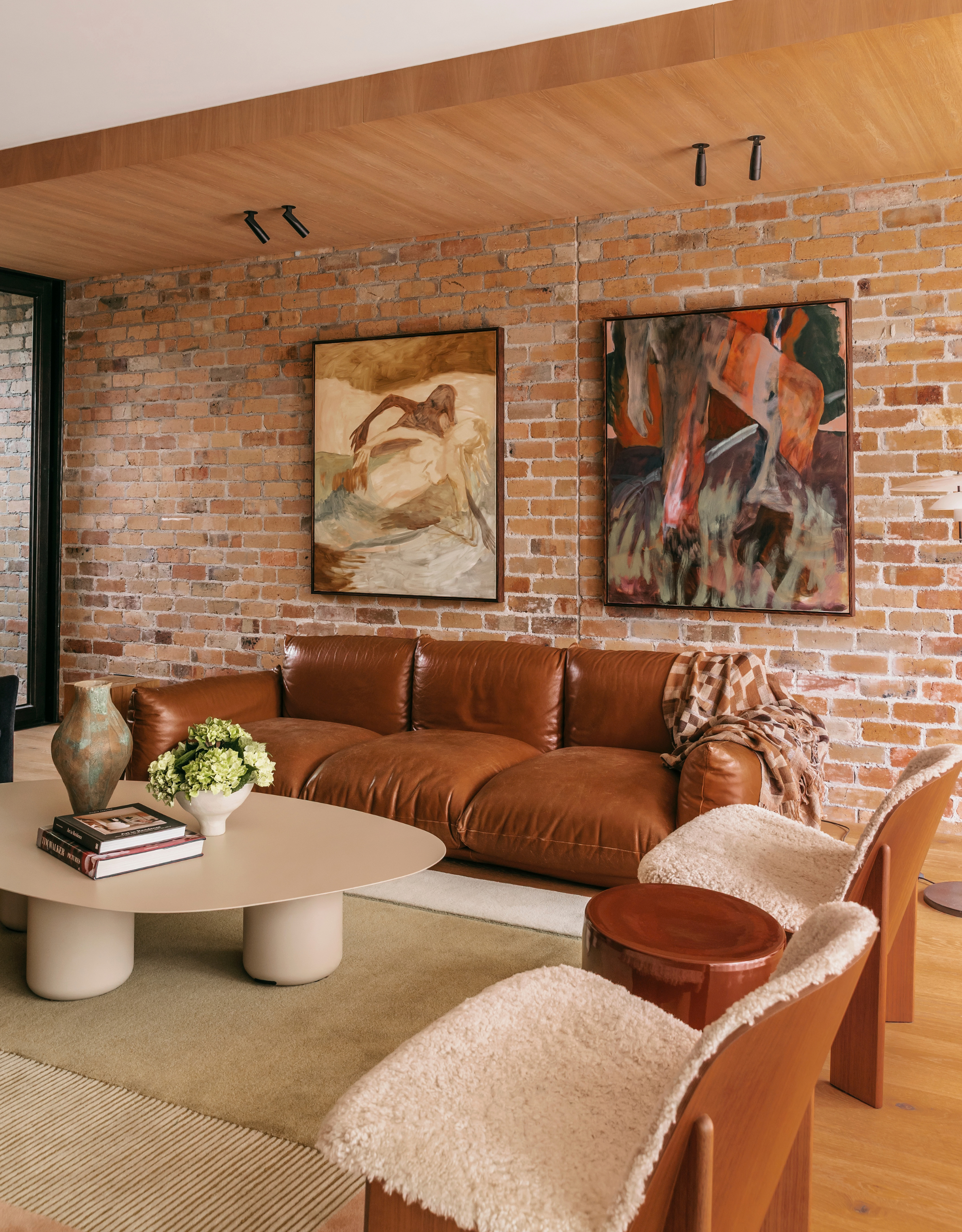 A brick living room with a leather sofa, a beige coffee table, sheepskin chairs, and paintings on the wall