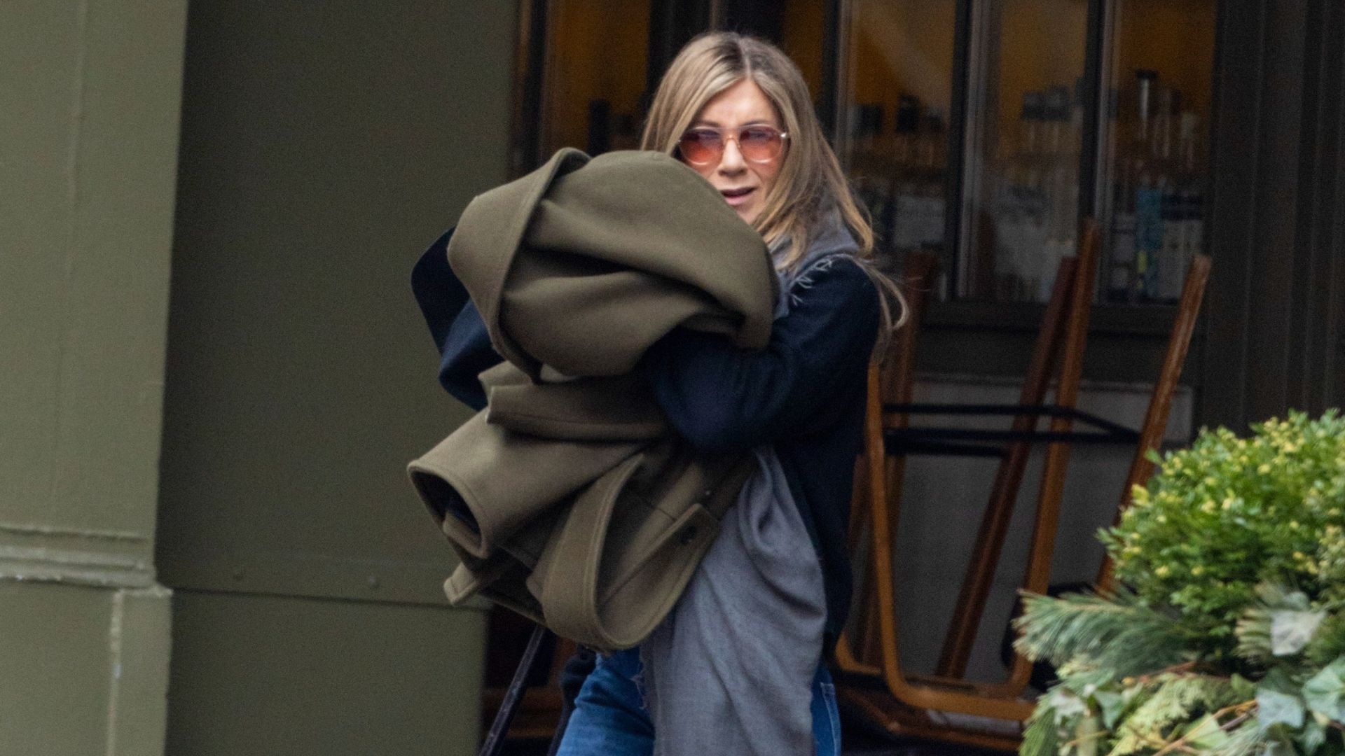 Jennifer Aniston wears puddle jeans with a navy knit and an olive green coat.