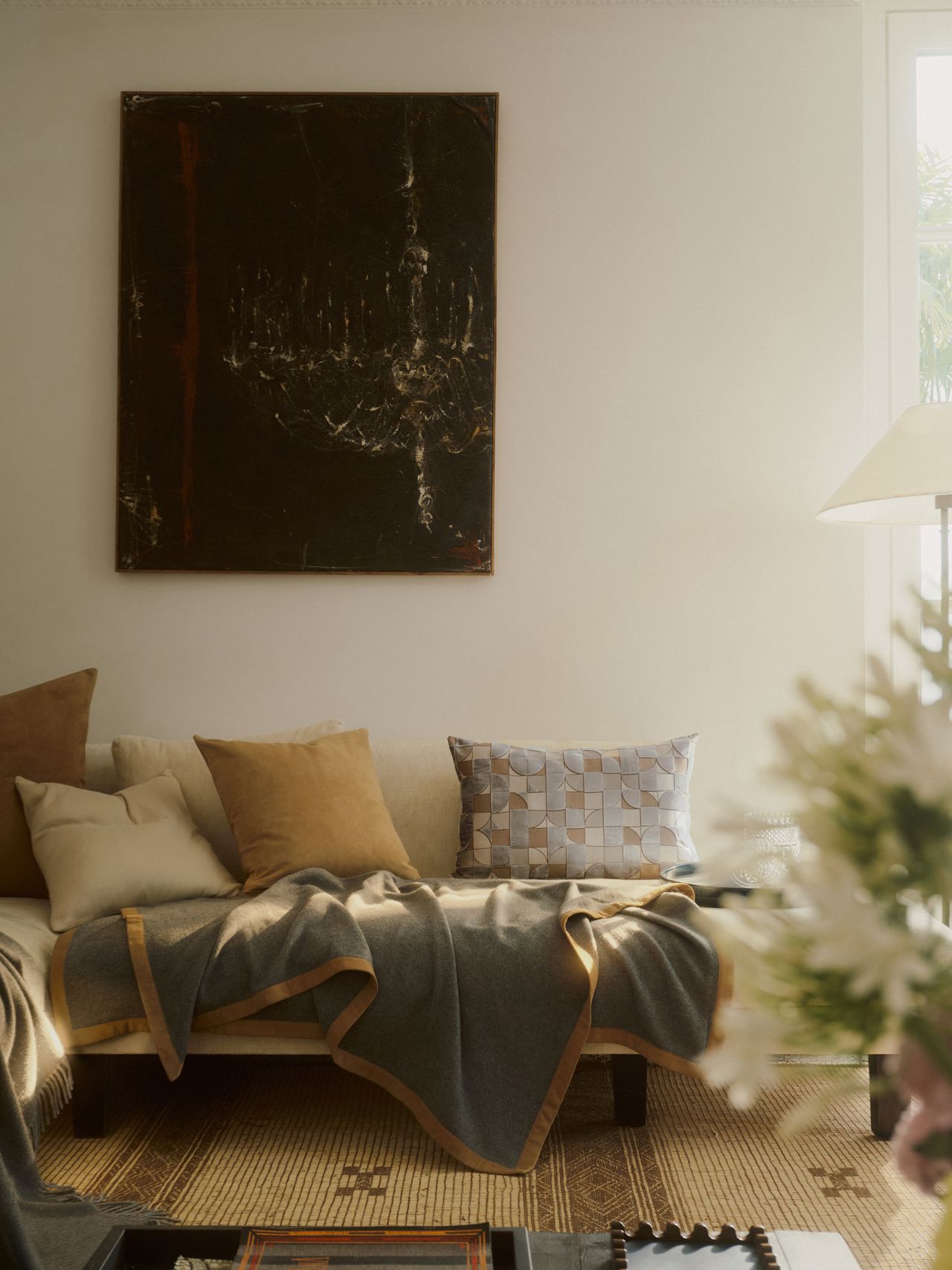 Image of an off-white room with a bench seat that has throw pillows and blankets on it. There is a black painting on the wall.